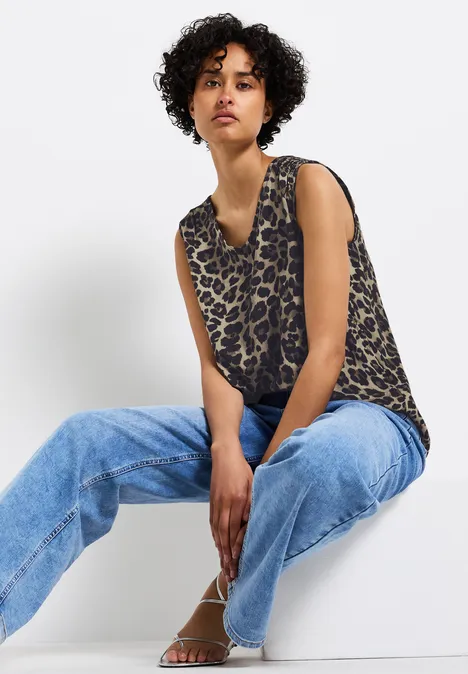 Frau mit Locken trägt Leoparden-Top und Jeans, sitzt entspannt auf weißem Hintergrund.